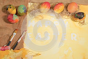 Easter colorful eggs with two painter's brushes,jute canvas,arranged on yellow painted watercolor paper.