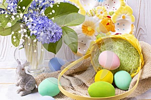 Easter colorful eggs in a wicker basket and a rabbit figurine and a spring bouquet in a vase and a beautiful ceramic plate on a