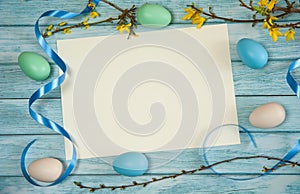 A blank sheet of paper, branches of forsythia with yellow flowers, ribbons and Easter eggs on a light blue wooden background.