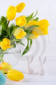 Easter composition with bouquet of yellow tulip flowers in glass vase and two white ceramic rabbits on white