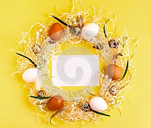 Easter creative concept. Wreath of white, brown chicken eggs, quail eggs, flowers and empty greeting card on a yellow background.