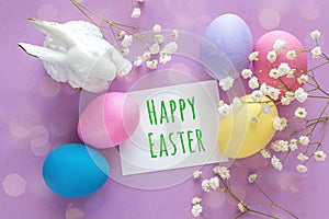 Easter decoration with message Happy Easter, eggs, rabbit and white flowers on a purple background.