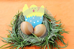 Easter decoration: yellow eggs and hand made hatched chicken in eggshell in green grass twigs nest on orange background