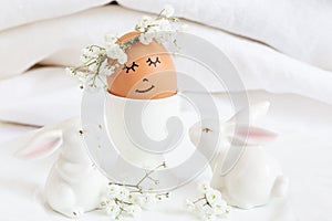 Easter egg with cute face in floral wreath crowns in egg cup with two white easter bunnies