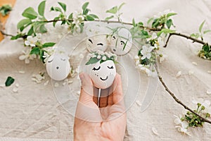 Easter egg with drawn cute face in floral wreath in person hand on background of eggs and petals