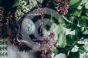 easter egg in nest with floral and chick ornaments on rustic background with lilac flowers. top view. space for text. happy
