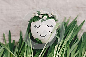Easter egg with sleeping face in floral wreath in grass on rustic linen  background. Happy Easter