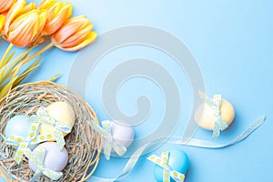 Easter eggs basket. Colorful egg with tape ribbon, spring tulips, white feathers on pastel blue background in Happy Easter