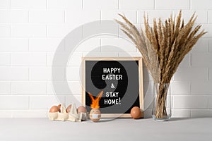 Easter eggs, and black felt letter board with slogan - Happy Easter and Stay Home on white brick wall background