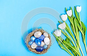 Easter eggs of blue flowers in a decorative nest of straw and a bouquet of white tulips on a blue background, copy space, flat la