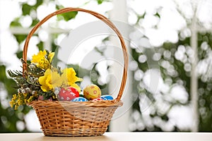 Easter eggs and bouquet in wicker basket on table against window, space for text