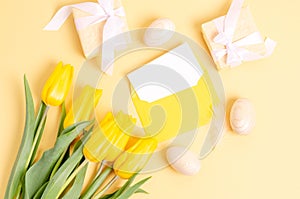Easter eggs, gift boxes, an empty postcard in an envelope and tulips on a yellow background
