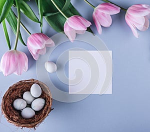 Easter eggs in nest, pink tulips flowers and sheet of paper over light blue background. Greeting card for Happy Easter. Flat lay