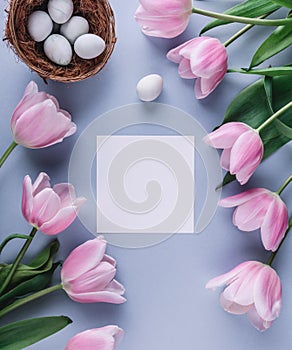 Easter eggs in nest, pink tulips flowers and sheet of paper over light blue background. Greeting card for Happy Easter. Flat lay,