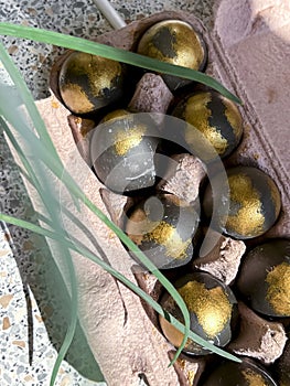 Easter eggs painted in black and gold are lying in a tray next to green leaves of grass.