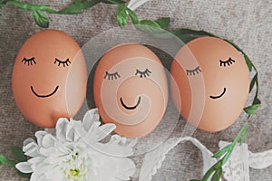 easter eggs with painted faces and smiles on a light beige background with white flowers and a green ribbon from plants, spring