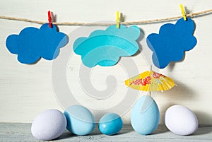 Easter eggs in pastel colors, yellow rice paper umbrella and paper clouds on a clothespins, on white wooden background
