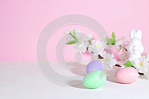 Easter eggs, pink, green and lilac, on a concrete white table, next to bunnie and flowers, on a pink background. Pastel shades