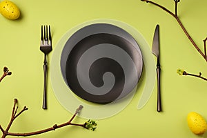 Easter festive table setting. Black plate, table knife and fork on a green background. Branches with leaves and yellow eggs. An