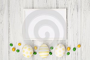 Easter frame with colorful eggs, yellow and green candies and copy space, flat lay on the white wooden table. Template, mockup