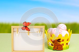 Easter greeting card template. Colorful Easter eggs in a basket and an empty wooden frame with a butterfly on table over natural