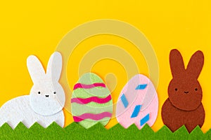 Easter holiday concept. Cut out of felt applications of two eggs and white and brown rabbits on the grass. Yellow background. Copy