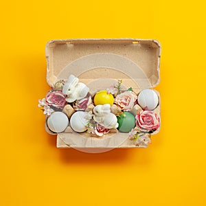 Easter holiday concept with cute handmade colorful eggs, egg tray, bunny and flowers on yellow background. Flat lay concept, top