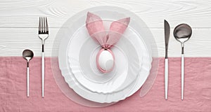 Easter holiday table setting with plate and cutlery, invitation concept. White wooden background. Bunny made of pink linen napkin