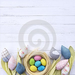 Easter nest with colorful eggs surrounded by fabric tulips flowers on a background of white wooden boards. A beautiful spring