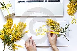 Easter spring flat lay with yellow flowers, laptop and a cup of coffee.Woman`s hand is writing text in notepad.