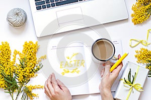 Easter spring flat lay with yellow flowers, laptop and a cup of coffee.Woman`s hand is writing text in notepad.