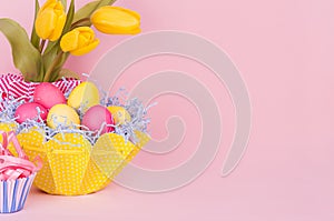 Easter spring home decor of yellow tulips, painted eggs, cupcake on pastel soft pink background.