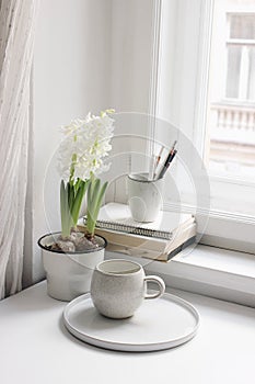 Easter spring still life. Cup of coffee, book and blank diary near window sill. White hyacinth in flower pot. Pencils in