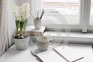 Easter spring still life. Cup of coffee, book and blank open diary mockup near window sill. White hyacinth in flower pot