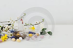 Easter still life. Colorful easter chocolate eggs in nest, spring flowers, chicken figurine composition on white wooden table.