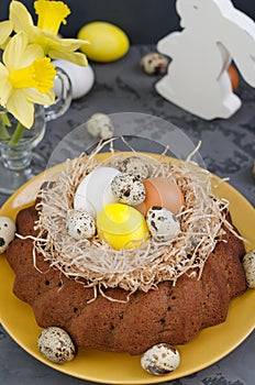 Easter still life, Easter cake with dyed eggs in a nest, daffodils in a cup on a concrete background