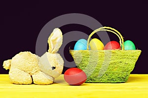 Easter symbols composition. Toy bunny and painted Easter eggs in different colours standing on yellow wooden table