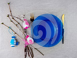 Easter table setting empty blue glass plate fork knife with tree branches linen cloth top view. Festive menu dish place