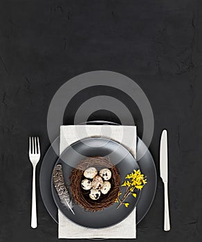 Easter table setting with a nest of quail eggs , a feather, a napkin and yellow flowers on a black background. Vertical