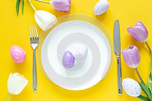 Easter table setting. White plate, decorative easter eggs, tulips and silver cutlery on yellow background