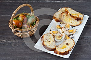 Easter wicker basket with colored eggs and sliced Easter bread in white plate on grey wooden board.