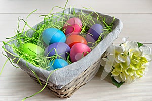 Easter wicker basket with colored eggs and a white flower on wooden board.