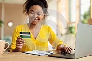 Easy payment and cashback concept. Smiling black lady doing online shopping at home using laptop and credit card