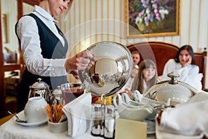 Eat and Drink. Caucasian parents and two kids waiting for breakfast delivered by waitress in unifrom in luxurious hotel