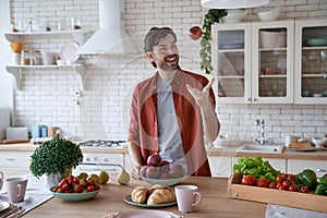 Eating apple every morning. Young happy bearded man in casual clothes throwing up a red apple and smiling while standing