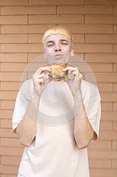 A eating Caucasian man who bitting and holding a burger, wearing a beige T-shirt