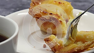 Eating with a dessert fork a fresh and hot crispy puff pastry pie with egg custard on a white plate at breakfast in a