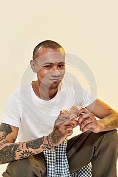 Eating food. A dark-skinned male with tattoos eating a hambuger with his mouth closed wearing a white t-shirt