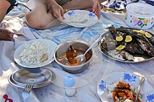 Eating fresh caught fish prepared into stew and grilled meat, detail to dishes with seafood and rice served on a blanket at the