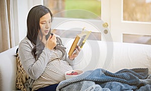 Eating healthy just became twice as important. an attractive young pregnant woman eating fruit and reading a book while